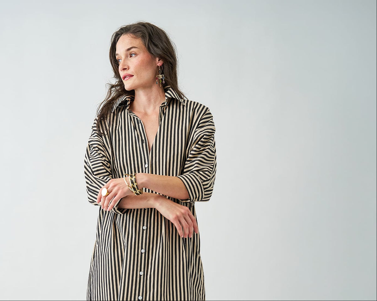 A woman wearing a black and beige vertically striped button-down shirt dress, standing against a light gray studio background.