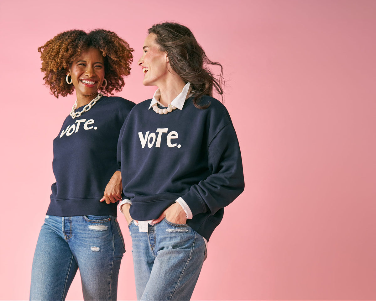 Two women wearing matching navy blue crewneck sweatshirts with the white text 'vote.' printed on the chest, standing against a solid pink studio background.